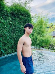 a shirtless man standing in front of a swimming pool next to a lush green bush