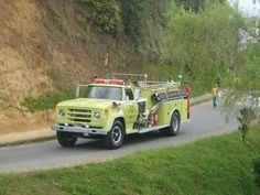 a yellow fire truck driving down a road next to a lush green hillside with trees