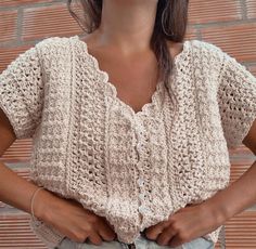 a woman standing in front of a brick wall with her hands on her hips and wearing a white crocheted top