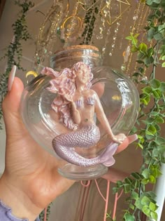 a hand holding a glass ball with a mermaid figure in it and greenery hanging from the ceiling