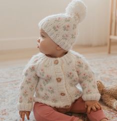 a baby sitting on the floor wearing a white knitted hat and sweater with flowers