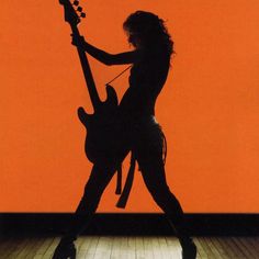 a woman holding a guitar in front of an orange wall with her hands on the strings