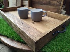 two coffee mugs sitting on top of a wooden tray in the middle of some grass