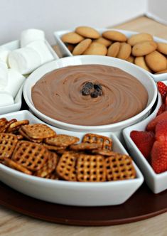 an assortment of snacks arranged in bowls on a wooden platter with marshmallows, strawberries and pretzels