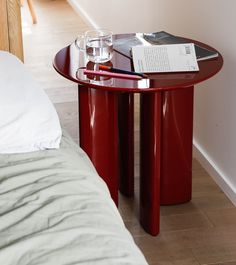 a red table with a book, glass and pen on it next to a bed