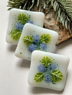 three square glass pieces with blue and green flowers on them next to a pine branch