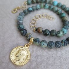 "A gold plated reproduction of a double-sided ancient Greek coin is the striking focal point of this African turquoise beaded necklace. The variegated blue-green coloring of the stones is the reason they are called \"turquoise\", but African turquoise is actually a jasper found in Africa. The gemstones have been finished to a velvety soft, matte patina to add to the necklace's appearance of an ancient and rare archeological find. A gold extender chain completes the necklace and allows it to be a Gold Turquoise Necklace With Round Beads And Natural Stones, Gold Turquoise Necklace With Gemstone Beads For Spiritual Style, Gold Turquoise Necklace With Round Gemstone Beads, Spiritual Gold Turquoise Necklace With Gemstone Beads, Handmade Gold Turquoise Necklace With Round Beads, Gold Turquoise Necklace With Round Beads For Gift, Gold Turquoise Necklace With Round Beads As A Gift, Ancient Style Gemstone Beads Jewelry For Gift, Ancient Style Gemstone Beads Jewelry Gift