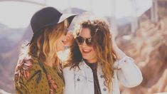 two women standing next to each other near the mountains and bridge in the background, both wearing sun glasses