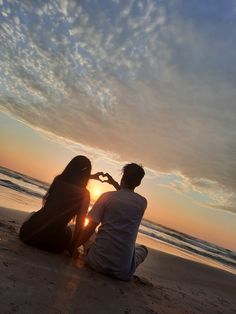 a man and woman sitting on the beach at sunset making a heart shape with their hands