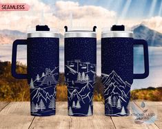 three travel mugs on a wooden table with mountains and lake in the background, one is blue