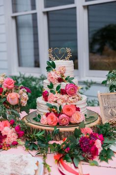 a wedding cake with pink flowers and greenery on a table in front of a window