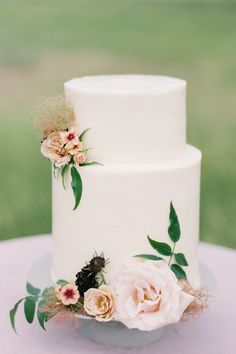 a white wedding cake with pink flowers on top and greenery around the edges, sitting on a table outside