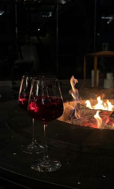 two glasses of red wine are sitting in front of an open fire pit at night