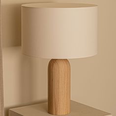 a white lamp sitting on top of a wooden table next to a beige wall with a curtain