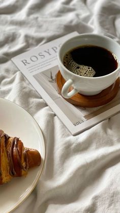 a pastry on a plate next to a cup of coffee and magazine laying on the bed