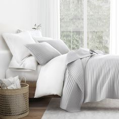 a bed with white sheets and pillows next to a basket on the floor in front of a window