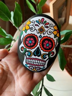 a hand holding a rock with a colorful sugar skull on it's face and green leaves in the background