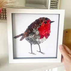 a bird made out of buttons sitting in a white frame on top of a table