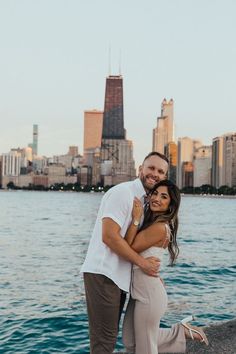 a man and woman standing next to each other near the water in front of a city skyline