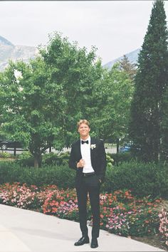 a man in a tuxedo standing on a sidewalk with trees and flowers behind him