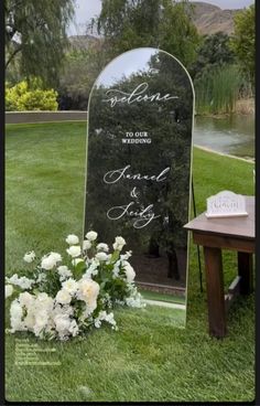 a wedding sign in the grass next to flowers and a table with a mirror on it