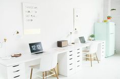 a white desk with two laptops on it and a refrigerator in the back ground