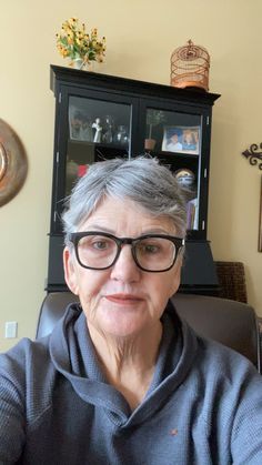 an older woman wearing glasses sitting in front of a book shelf and looking at the camera