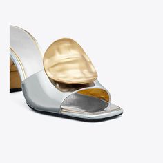 Our Patos sandal is defined by mismatched metal discs, evocative of ’60s glamour. Crafted in metallic leather, the sculptural silhouette features a slanted heel that gives it a chic, distorted look. A cushioned insole ensures day-to-night comfort. Modern Evening Mules With Single Toe Strap, Metallic Sandals With Sculpted Heel For Formal Occasions, Cocktail Mules With Sculpted Heel And Single Toe Strap, Formal Metallic Sandals With Sculpted Heel, Luxury Silver Open Toe Mules, Modern Metallic Sandals For Formal Occasions, Luxury Silver Mules For Party, Modern Metallic Mules For Evening, Modern Metallic Mules For Formal Occasions