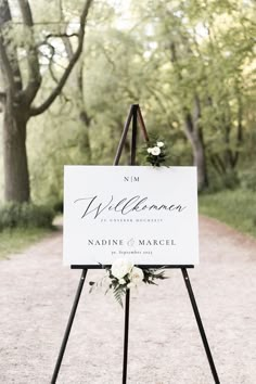 a welcome sign with flowers on it in the middle of a dirt road surrounded by trees