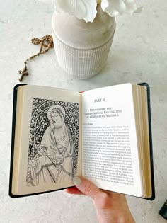 a person holding an open book in front of a vase with flowers on the table