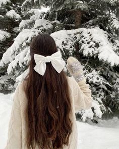 a woman with long hair wearing a white bow in front of snow covered pine trees