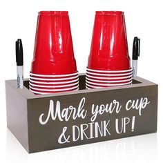 two red cups are in a wooden box with writing on the front and bottom, along with black marker pens