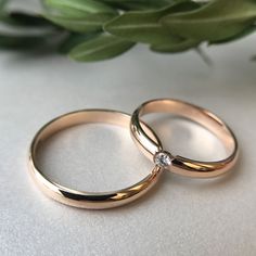 two gold wedding rings sitting next to each other on top of a white table with greenery