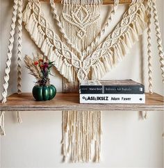 two books are sitting on a shelf next to a macrame wall hanging and a vase with flowers