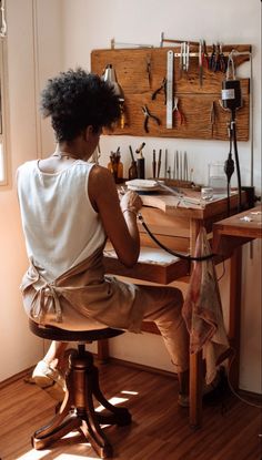 a woman sitting at a wooden desk working on an object with tools hanging on the wall behind her