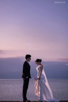 a man and woman standing next to each other near the ocean at sunset or dawn