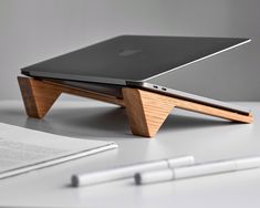 an apple laptop on a wooden stand next to a notepad and pen, with a notebook in the background