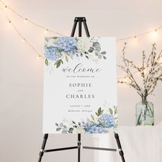 a welcome sign with blue hydrangeas and greenery sits on an easel