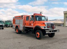 an orange fire truck parked in a parking lot with other trucks behind it and snow covered mountains in the background