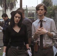 a man and woman walking down the street with other people behind them, one wearing a tie