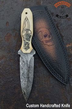 a knife with a skull on it and a leather sheath in the back ground next to another knife