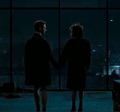 two people standing next to each other in front of a window with city lights behind them