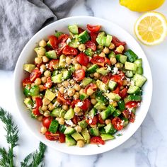 a white bowl filled with cucumber and chickpea salad next to lemon wedges