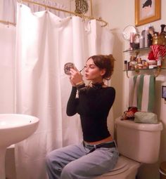 a woman sitting on top of a toilet next to a bathroom sink holding a mirror