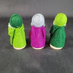 three small dolls in different colors and sizes on a gray surface, one is wearing a green jacket