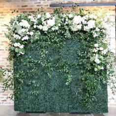 an artificial green wall with white flowers and greenery on the top, in front of a brick wall