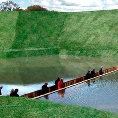 several people are standing on a long wooden bridge over a body of water with green grass in the background