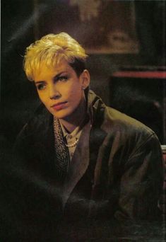 a young man with blonde hair wearing a black jacket and tie sitting in front of a piano