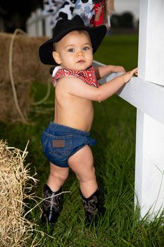 a baby wearing cowboy boots and a bandana is leaning against a white wall with hay bales in the background