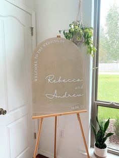 an easel with a wedding sign on it in front of a window and potted plant